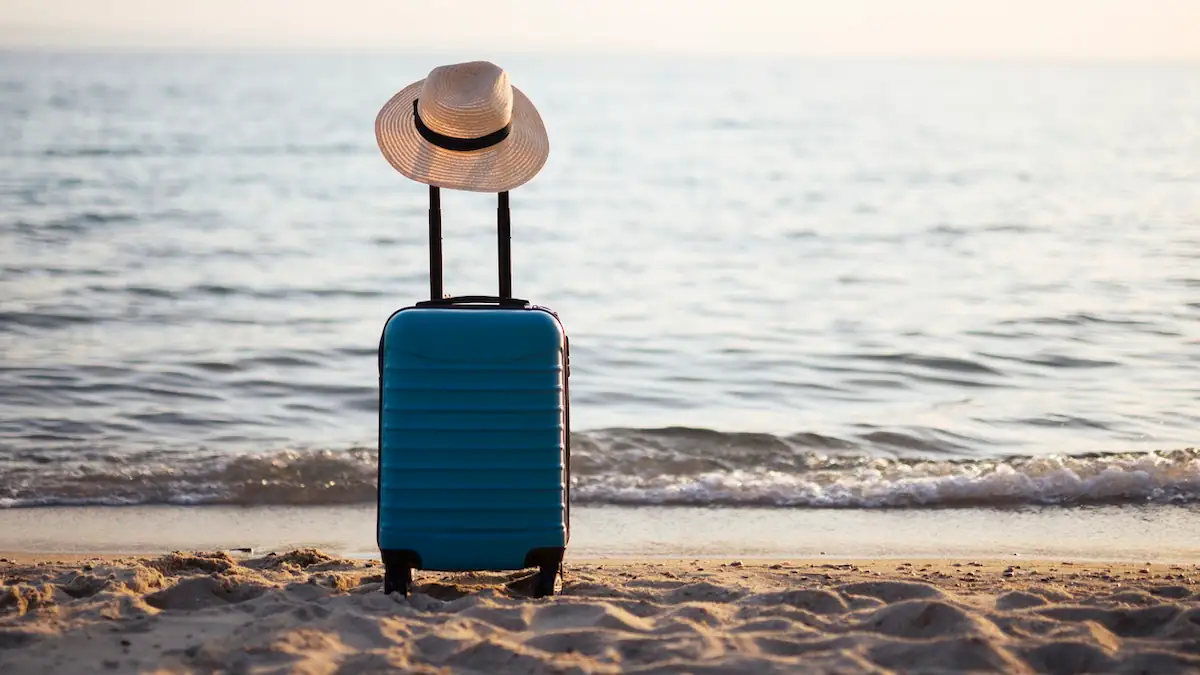 Handgepäck: Kleiner blauer Koffer mit Sonnenhut am Strand