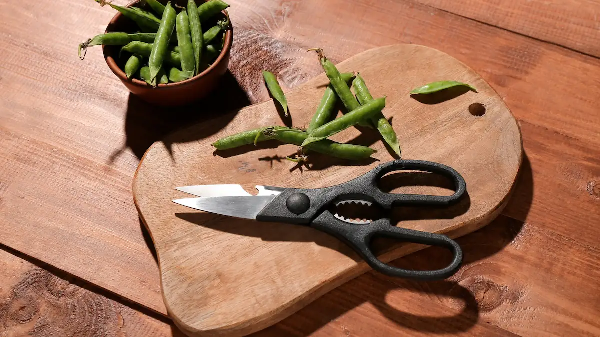 Küchenschere mit Metallzacken am Griff liegt neben Bohnen auf einem Holzbrett