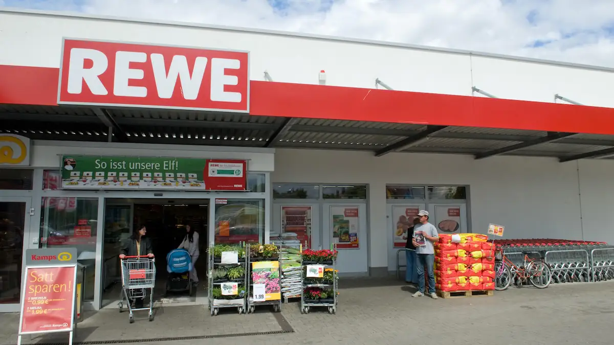 Rewe-Markt von vorne mit einigen Ainkäufern; vor dem Markt ist unter anderem Grillkohle in Säcken zu sehen