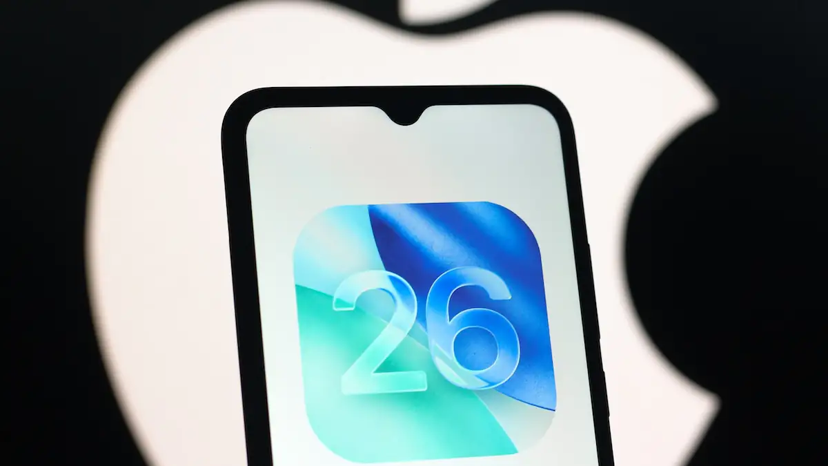 iOS 26: Abbildung des OS-Logos auf einem iPhone, im Hintergrund ist ein verschwommenes weißes Apple-Logo auf schwarzem Grund