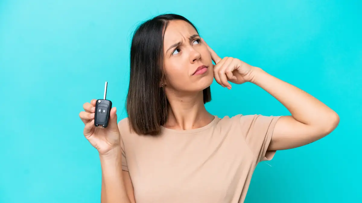 Junge Frau vor türkisfarbenem Hintergrund schaut nachdenklich nach oben, eine Hand am Kopf, in der anderen einen Autoschlüssel