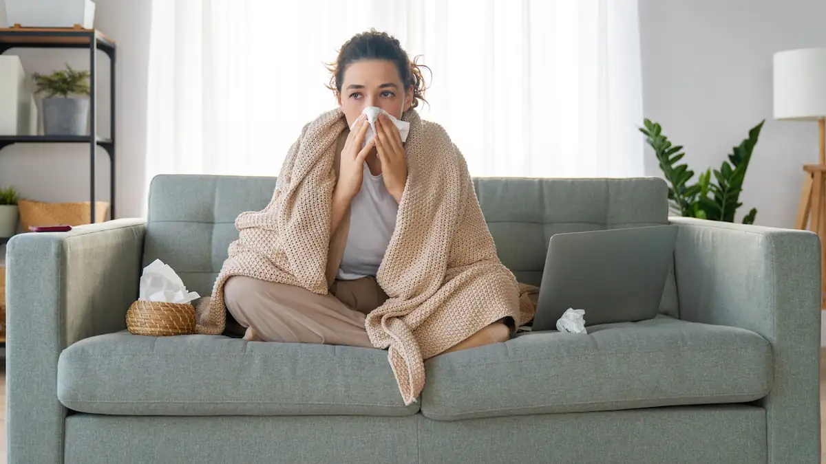 Frau sitzt mit Erkältung auf dem Sofa und putzt sich die Nase