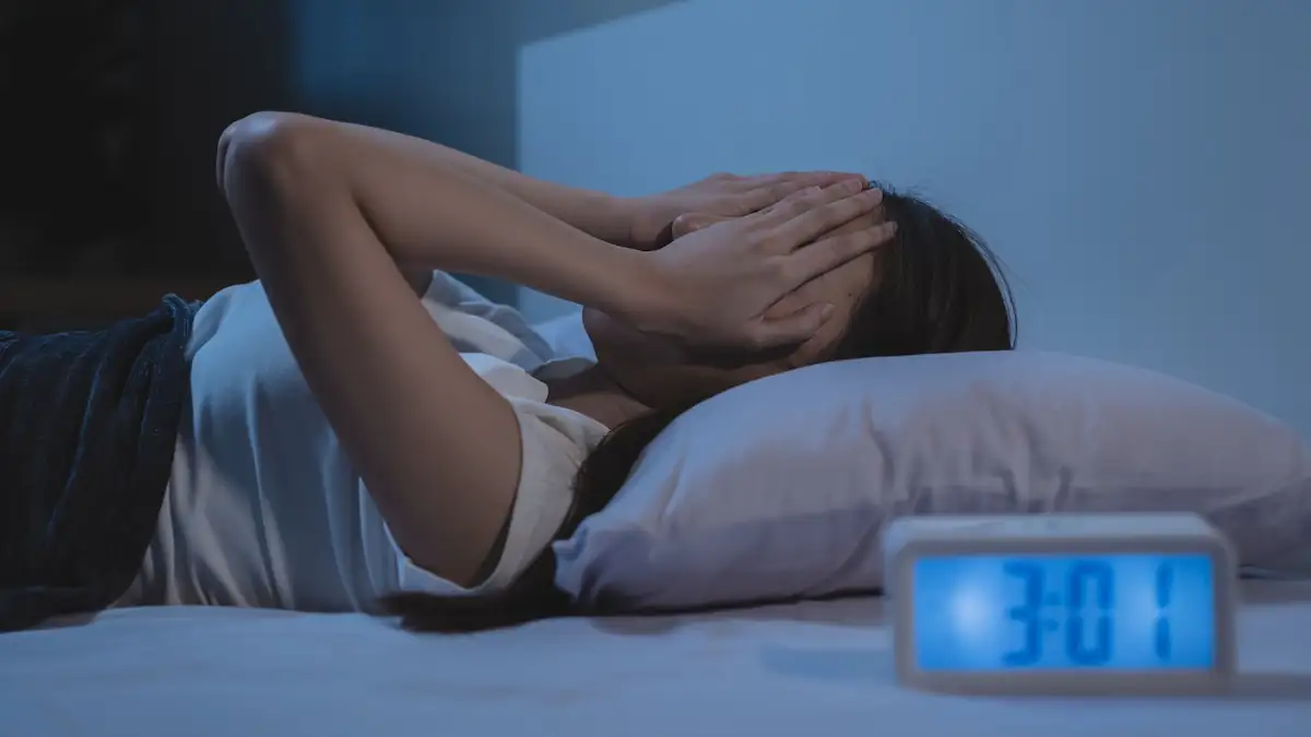 Wolfsstunde: Frau liegt schlaflos im Bett, Wecker im Vordergrund zeigt 03:01 Uhr nachts