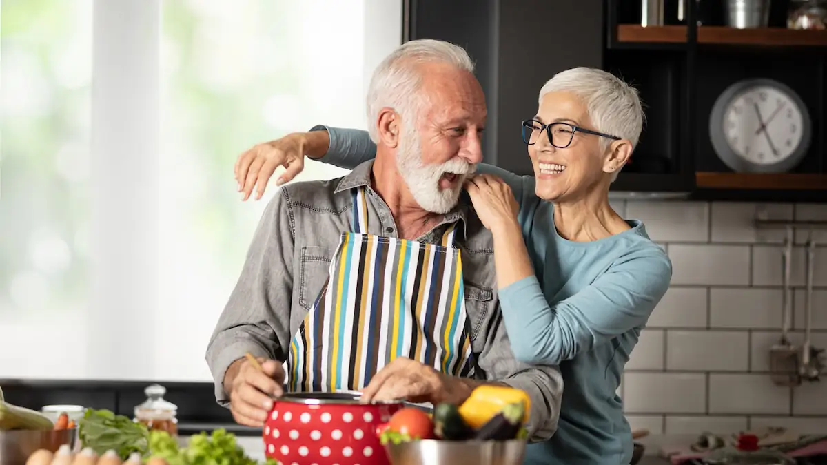 Zwei Personen in höherem Alter, ein Mann und eine Frau, stehen glücklich zusammen beim Kochen in der Küche