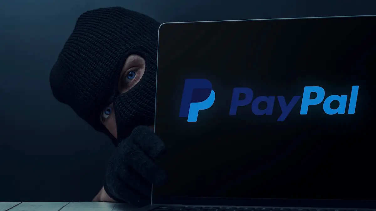 Maskierte Person schaut hinter einem Laptop hervor, auf dem ein PayPal-Logo zu sehen ist