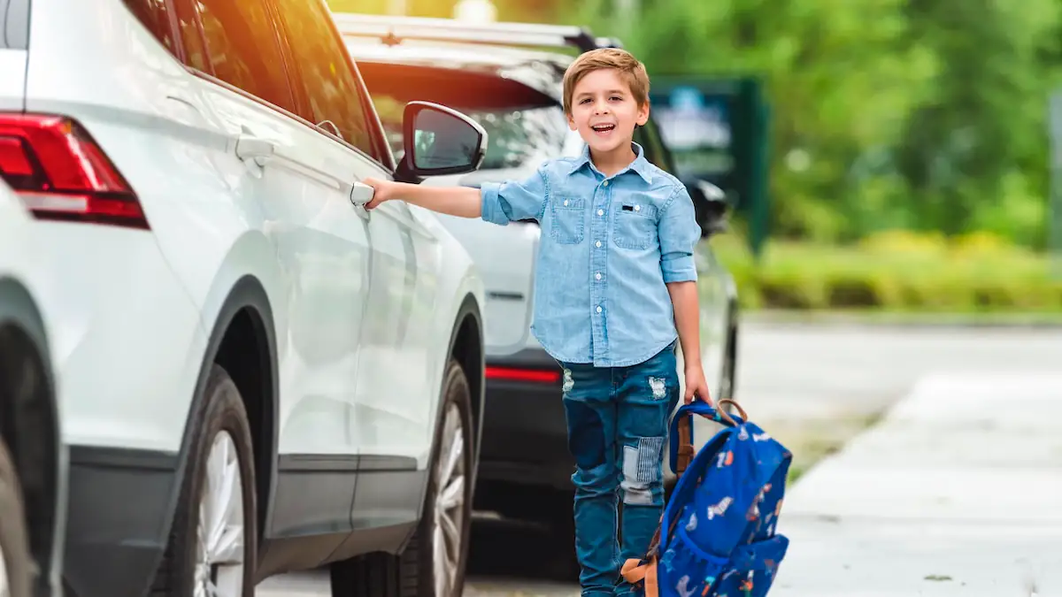 Elterntaxi: Kleiner Schuljunge steht mit seinem Ranzen neben einem Auto, die Hand im Griff