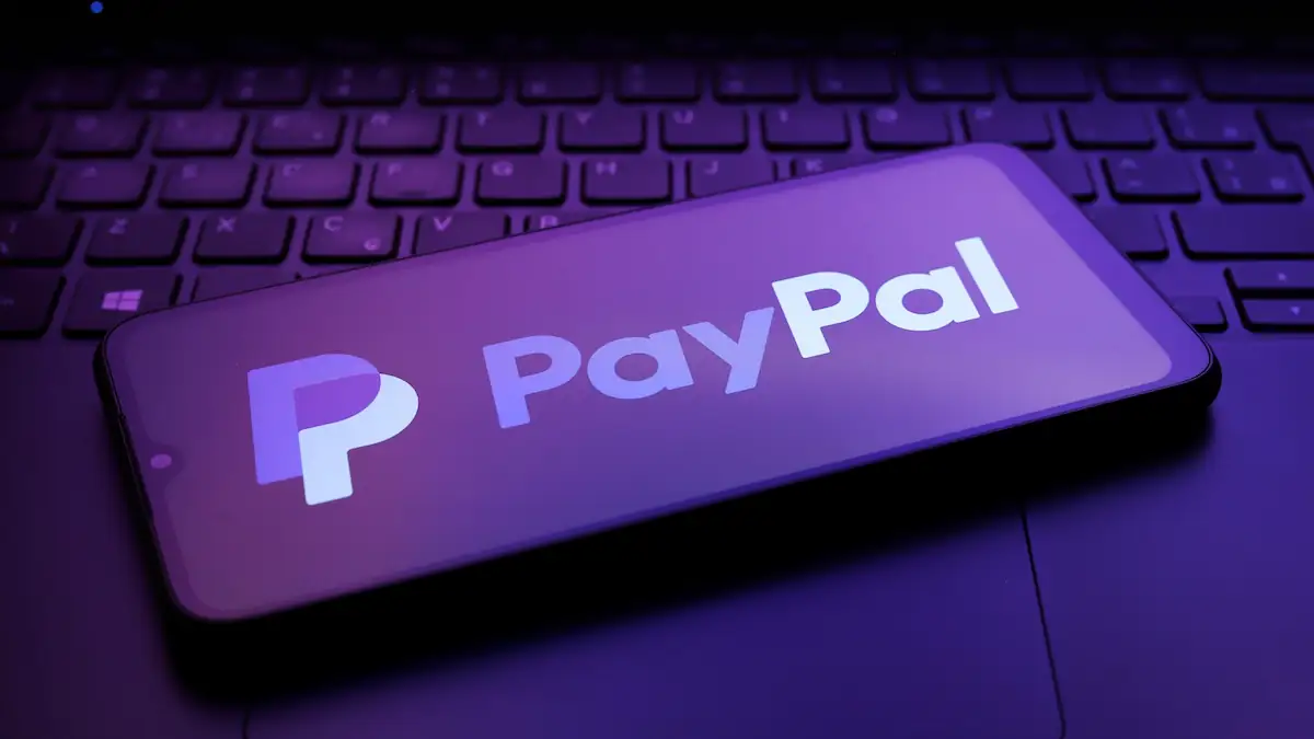 Smartphone mit PayPal-Logo auf dem Display liegt in lilafarbenem Licht auf einer Laptop-Tastatur