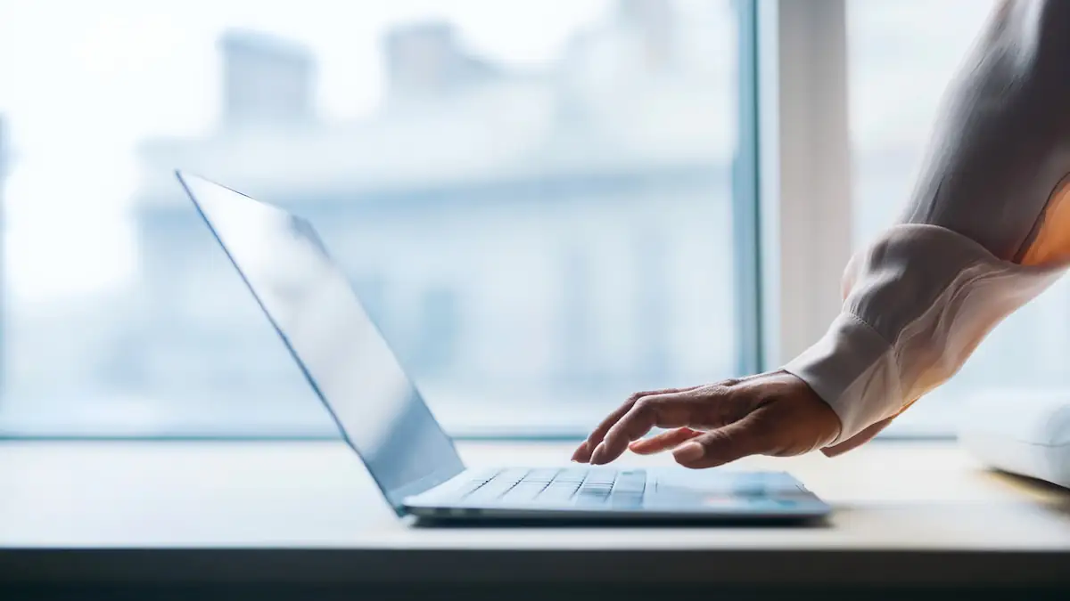 Aufgeklapptes Notebook vor einem Fenster, über dessen Tastatur eine Hand schwebt
