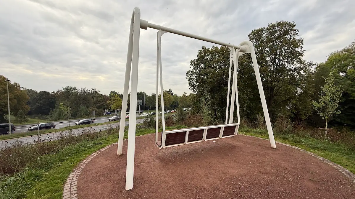 Eine Hollywood-Schaukel steht auf einem eigens aufgeschütteten Hügel mit Blick auf die vierspurige Bundesstraße 3 (B3), die direkt zur Autobahn A49 führt. Hinter ihr liegt der Park Schönfeld-Ost. Die Schaukel wird im sogenannten Schwarzbuch des Bunds der Steuerzahler Hessen genannt.