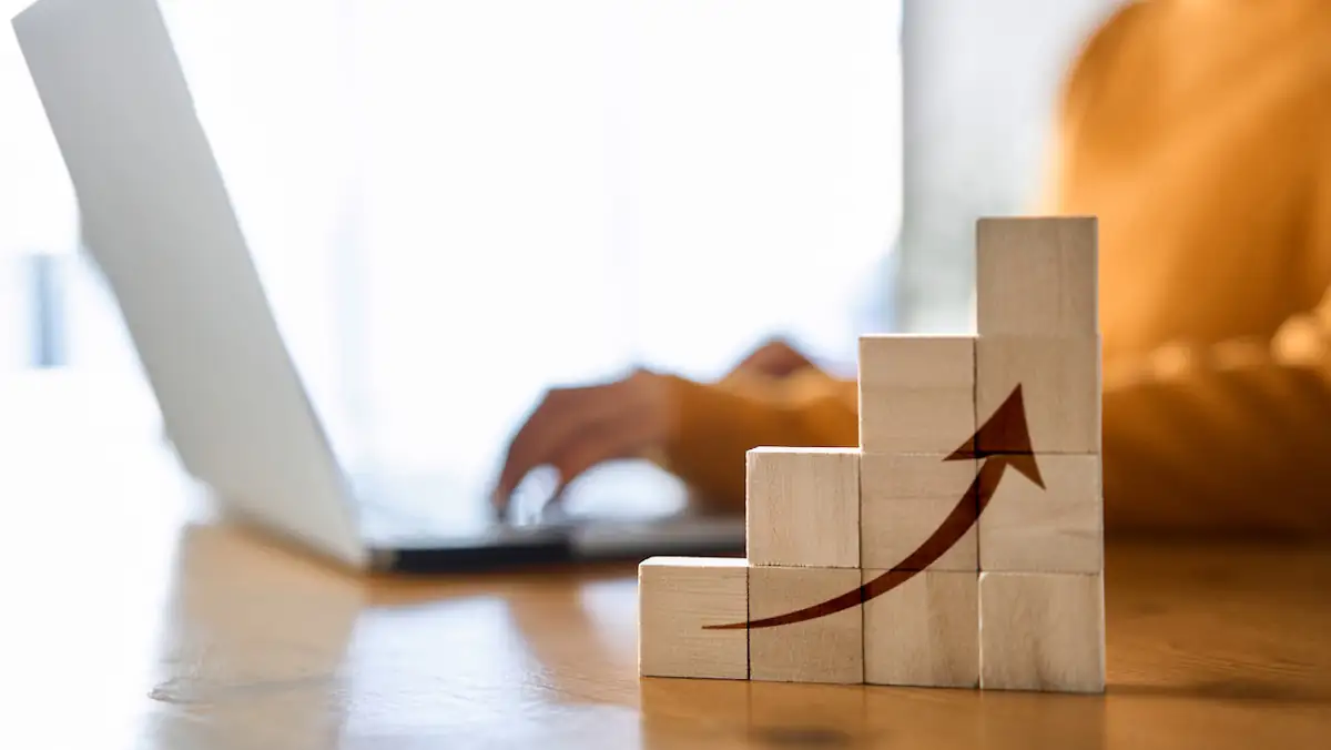 Symbolbild Nebenjob: Frau sitzt an Laptop, davor mit Bausteinen das Bild einer nach oben zeigenden Kurve mit Pfeil